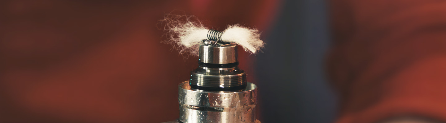 Eine Nahaufnahme eines Vape-Coils mit Watte. Der Coil ist aus Metall und wird von flauschiger, weißer Watte durchzogen, die etwas zerfasert ist. Das Setup befindet sich auf einem glänzenden Metallverdampfer, der leicht reflektiert. Der Hintergrund ist verschwommen und hat warme, rötlich-braune Farbtöne.