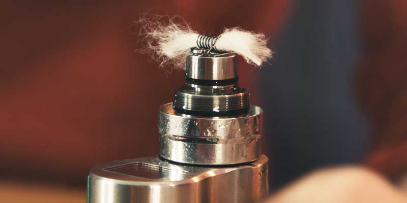 Eine Nahaufnahme eines Vape-Coils mit Watte. Der Coil ist aus Metall und wird von flauschiger, weißer Watte durchzogen, die etwas zerfasert ist. Das Setup befindet sich auf einem glänzenden Metallverdampfer, der leicht reflektiert. Der Hintergrund ist verschwommen und hat warme, rötlich-braune Farbtöne.