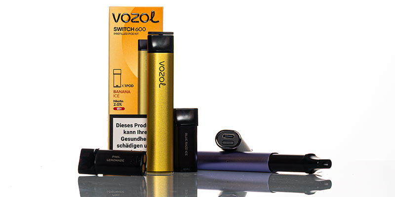 Mehrere Vozol Switch 600 Einweg-Vapes in verschiedenen Farben und Geschmacksrichtungen, darunter eine goldene Vape mit «Banana Ice»-Geschmack und austauschbare Pods. Die Verpackung mit Vozol-Logo und Gesundheitswarnung ist im Hintergrund sichtbar. Spiegelnde Oberfläche und weisse Hintergrundbeleuchtung. 	