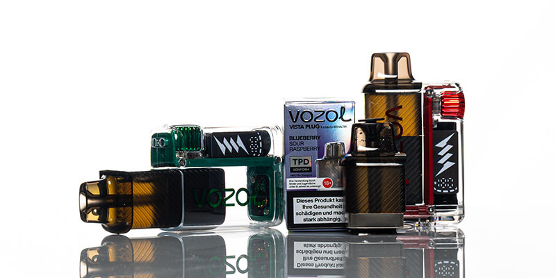 Mehrere Vozol Vista Plug Vapes mit transparentem Gehäuse, durch das die interne Elektronik sichtbar ist. Die Vapes sind in verschiedenen Farben gestaltet, darunter grün und schwarz-gold. Im Hintergrund steht eine Verpackung mit der Geschmacksrichtung «Blueberry Sour Raspberry» und einer Gesundheitswarnung. Spiegelnde Oberfläche mit weißem Hintergrund.