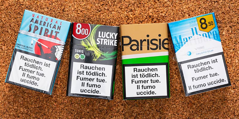 Vier Zigarettenpackungen unterschiedlicher Marken liegen auf einer Korkoberfläche. Auch hier ist der Warnhinweis in mehreren Sprachen prominent auf den Packungen abgedruckt.